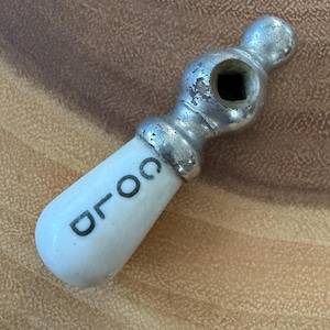 May include: A vintage faucet handle featuring a white ceramic lever with the word COLD printed in black lettering. The handle has a metallic silver-toned base with a square-shaped opening at the top, showing signs of wear and patina on the metal.