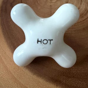 May include: A white, four-pronged ceramic faucet handle with a smooth, rounded cross shape. The center of the handle features the word HOT printed in clear, black, sans-serif capital letters, providing a functional label for the fixture.