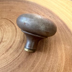 May include: A vintage metal door knob with a rounded, weathered grey surface and a tapered neck. The base of the knob features a small, circular brass-toned ring, showing signs of age and oxidation across its metallic, textured exterior finish.