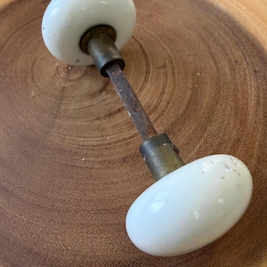 May include: A vintage door knob set featuring two smooth, round white porcelain handles connected by a central metal spindle. The hardware includes brass-toned collars at the base of each handle, showing signs of age and oxidation on the metal rod.
