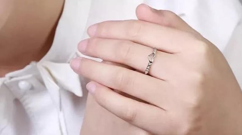 May include: A silver ring with a heart-shaped design. The ring features a delicate open heart at the top and small, round embellishments along the band. The ring is worn on a finger, showcasing its simple elegance.