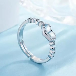 May include: A silver-toned ring with a heart-shaped design. The ring features a series of small, clear gemstones and a heart-shaped centerpiece. The ring is set against a light blue background.