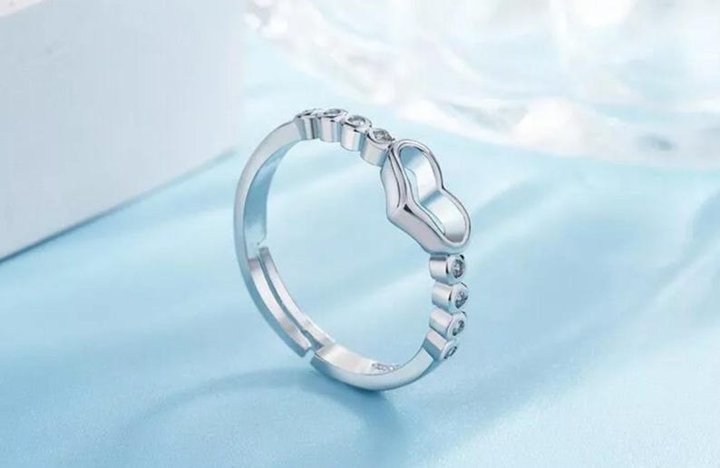 May include: A silver-toned ring with a heart-shaped design. The ring features a series of small, clear gemstones and a heart-shaped centerpiece. The ring is set against a light blue background.