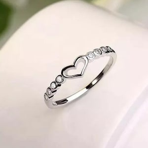May include: A silver ring with a heart-shaped cutout in the center. The ring is adorned with small, round, clear gemstones. The ring is set against a soft, white background, highlighting its delicate design.