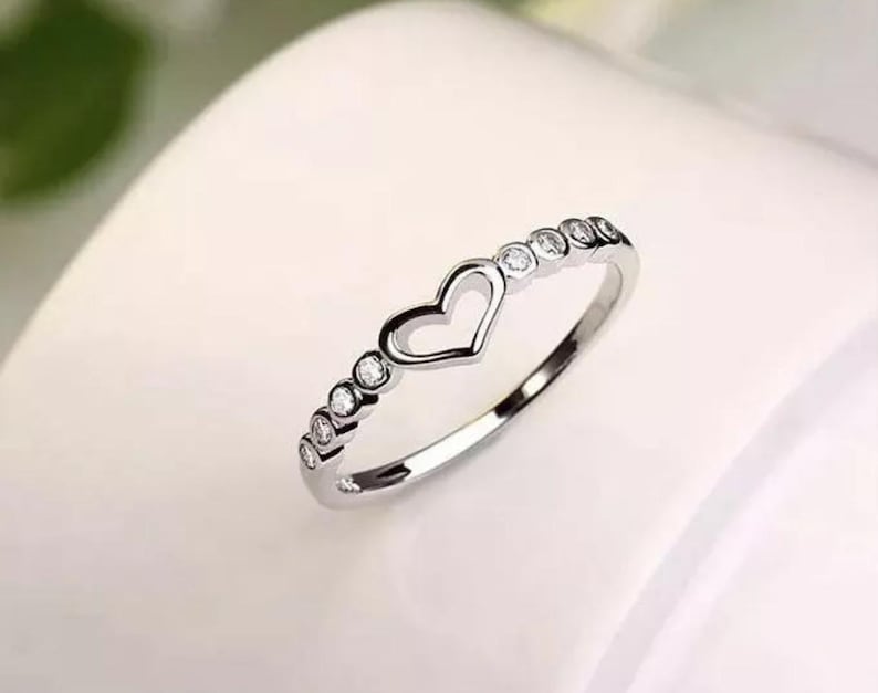 May include: A silver ring with a heart-shaped cutout in the center. The ring is adorned with small, round, clear gemstones. The ring is set against a soft, white background, highlighting its delicate design.