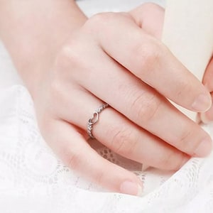 May include: A silver ring with a heart-shaped design. The ring features a small open heart in the center, surrounded by small, clear stones. The ring is worn on a finger, showcasing its delicate and romantic style.