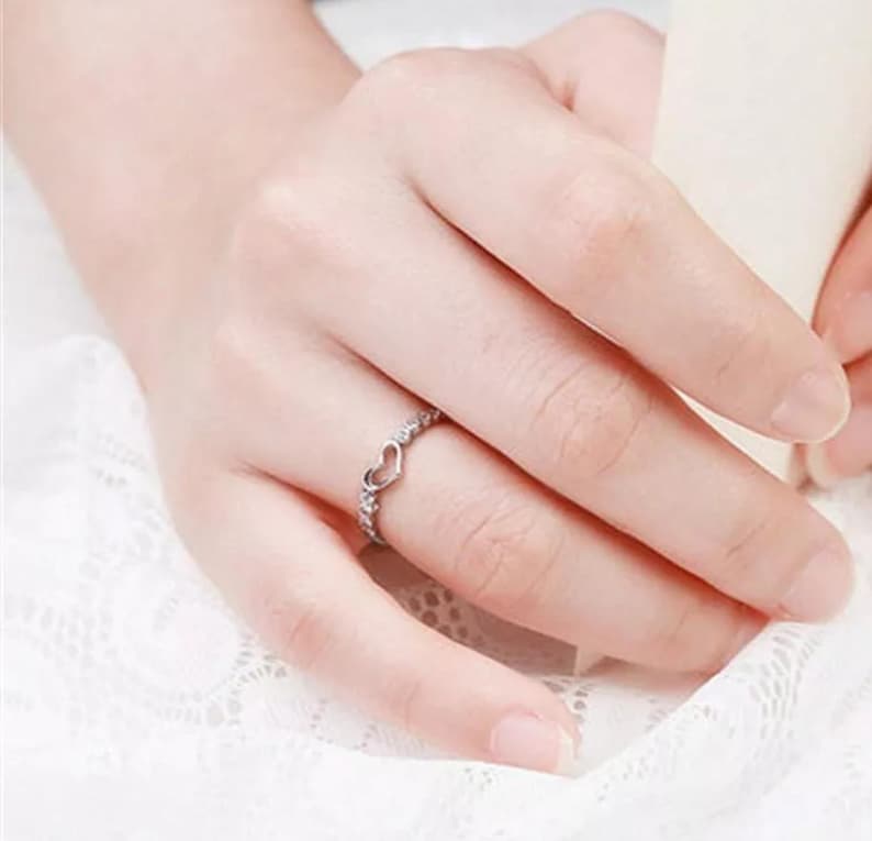 May include: A silver ring with a heart-shaped design. The ring features a small open heart in the center, surrounded by small, clear stones. The ring is worn on a finger, showcasing its delicate and romantic style.