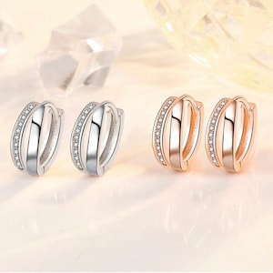 May include: Two pairs of small hoop earrings, one pair in silver tone and one pair in rose gold tone. Each earring features a multi-band design with one band encrusted with small clear stones and two smooth, polished bands alongside it.