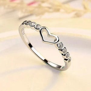 May include: A silver ring with a heart-shaped cutout in the center. The ring is adorned with small, round, clear gemstones. The band features additional gemstones, adding sparkle to the jewelry. The ring is set against a soft, blurred background.