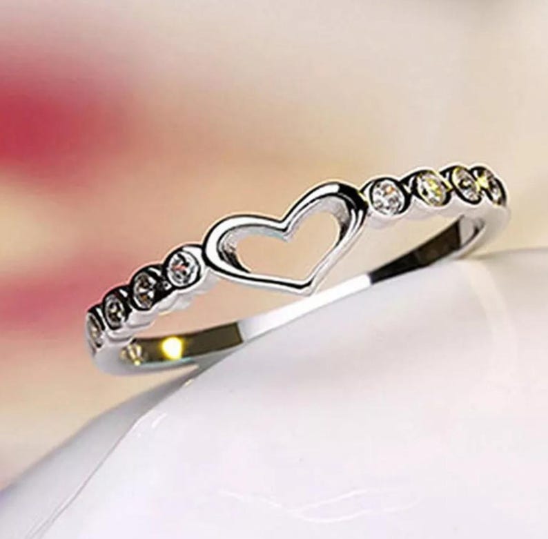 May include: A silver ring with a heart-shaped cutout in the center. The band is adorned with small, round, clear gemstones. The ring is displayed on a white surface with a soft pink and white background.