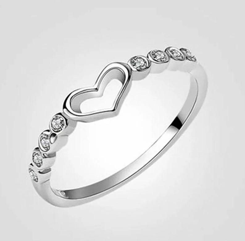 May include: A silver ring with a heart-shaped cutout and a row of small, clear gemstones. The ring has a polished finish and a simple, elegant design, perfect for everyday wear or special occasions.