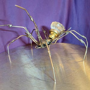 May include: A handcrafted metal spider sculpture with a clear glass knob head and brass accents. The spider's body is made of dark metal, and its legs are crafted from silver-colored wire. The sculpture is displayed on a silver surface against a purple backdrop.
