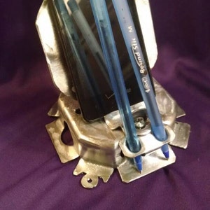May include: A silver-colored metal desk organizer holding a black smartphone and two blue BIC Round Stic pens. The organizer has a unique, industrial design and sits on a purple surface.