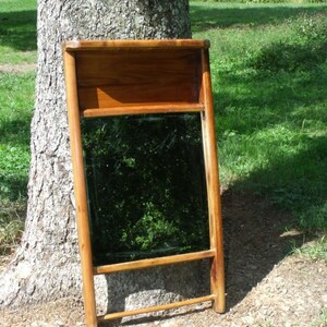 May include: A wooden framed mirror with a rectangular reflective surface. The frame is a warm brown color, and the mirror is leaning against a tree. The mirror has a shelf at the top and a horizontal bar at the bottom.
