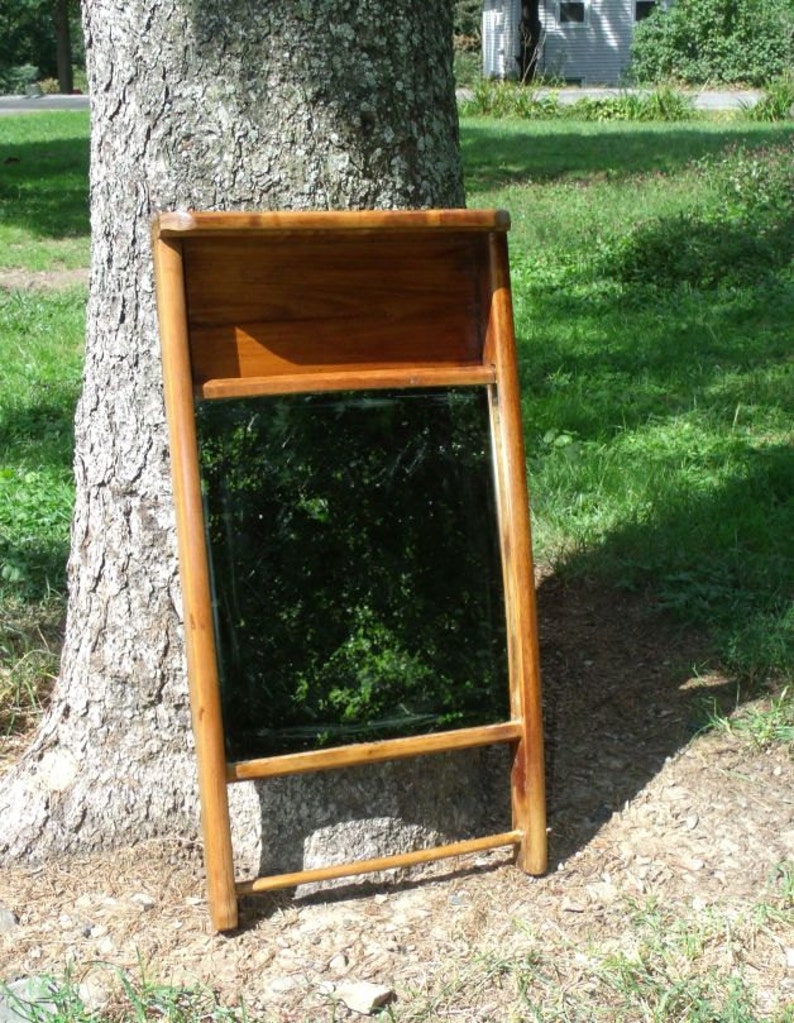 May include: A wooden framed mirror with a rectangular reflective surface. The frame is a warm brown color, and the mirror is leaning against a tree. The mirror has a shelf at the top and a horizontal bar at the bottom.
