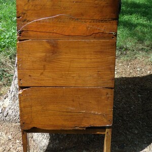May include: A weathered, wooden antique washstand with a warm, brown finish. The stand features a rectangular body constructed from horizontal planks, supported by four slender legs and a lower stretcher. The wood shows signs of age and wear.