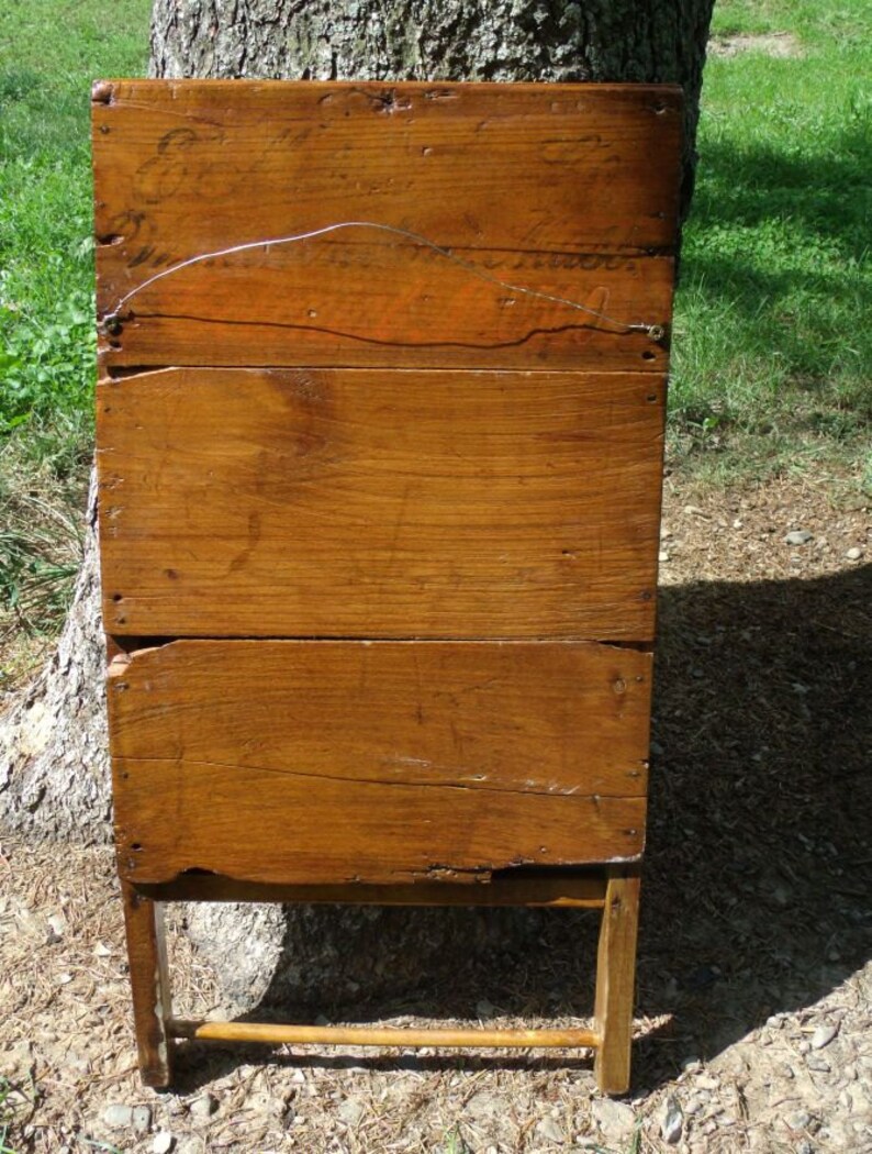 May include: A weathered, wooden antique washstand with a warm, brown finish. The stand features a rectangular body constructed from horizontal planks, supported by four slender legs and a lower stretcher. The wood shows signs of age and wear.