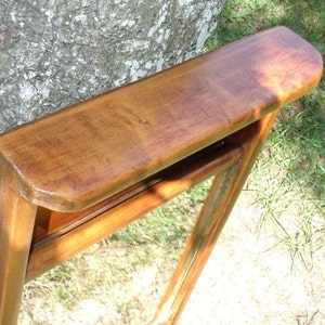 May include: A wooden mirror with a rectangular frame and a curved top. The frame is a warm brown color, and the mirror is angled, showing a reflection. The mirror is leaning against a tree trunk, with green grass in the background.