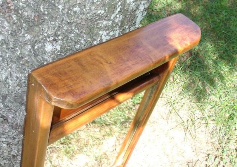 May include: A wooden mirror with a rectangular frame and a curved top. The frame is a warm brown color, and the mirror is angled, showing a reflection. The mirror is leaning against a tree trunk, with green grass in the background.