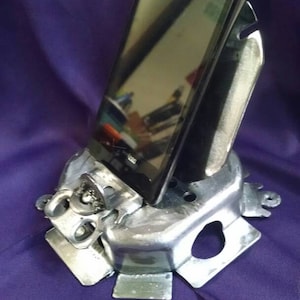 May include: A silver-colored, handmade phone stand made from repurposed metal components. An LG smartphone is placed in the stand, angled for viewing. The stand sits on a purple surface.