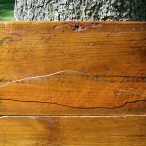 May include: A rectangular wooden sign with a warm, brown finish. The sign has visible wood grain and appears to be made of several planks joined together. Faint, aged lettering is visible on the surface, and a wire is strung across the back for hanging.