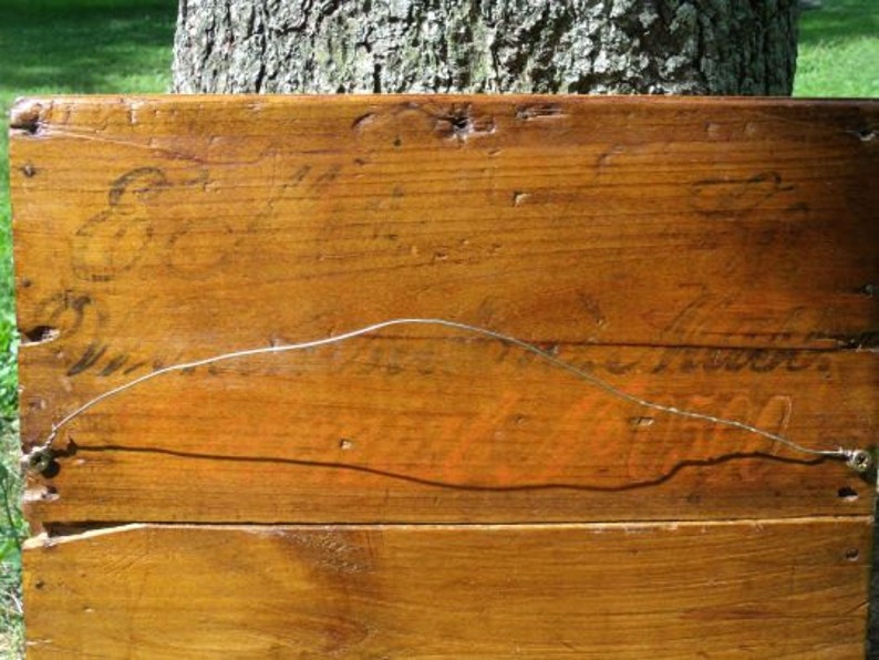 May include: A rectangular wooden sign with a warm, brown finish. The sign has visible wood grain and appears to be made of several planks joined together. Faint, aged lettering is visible on the surface, and a wire is strung across the back for hanging.