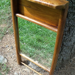 May include: A wooden mirror with a shelf, featuring a warm, brown finish. The mirror is rectangular, framed by vertical supports and horizontal bars. The shelf is at the top, and the mirror reflects the green grass.