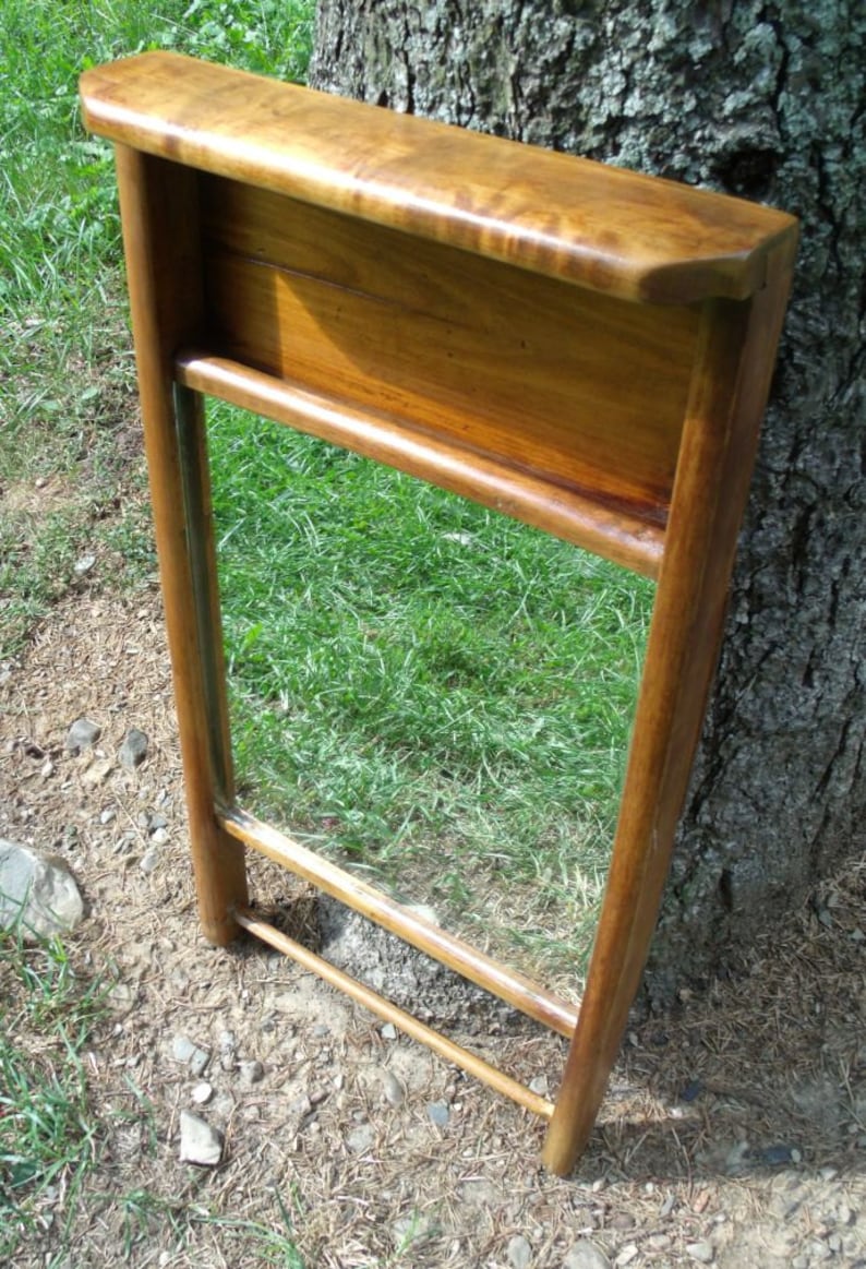 May include: A wooden mirror with a shelf, featuring a warm, brown finish. The mirror is rectangular, framed by vertical supports and horizontal bars. The shelf is at the top, and the mirror reflects the green grass.