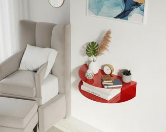 May include: A semi-circular red wall-mounted shelf holding a small white vase with a green plant, a round clock, a stack of books, and a small potted succulent. The shelf is positioned on a white wall next to a light gray armchair and ottoman.
