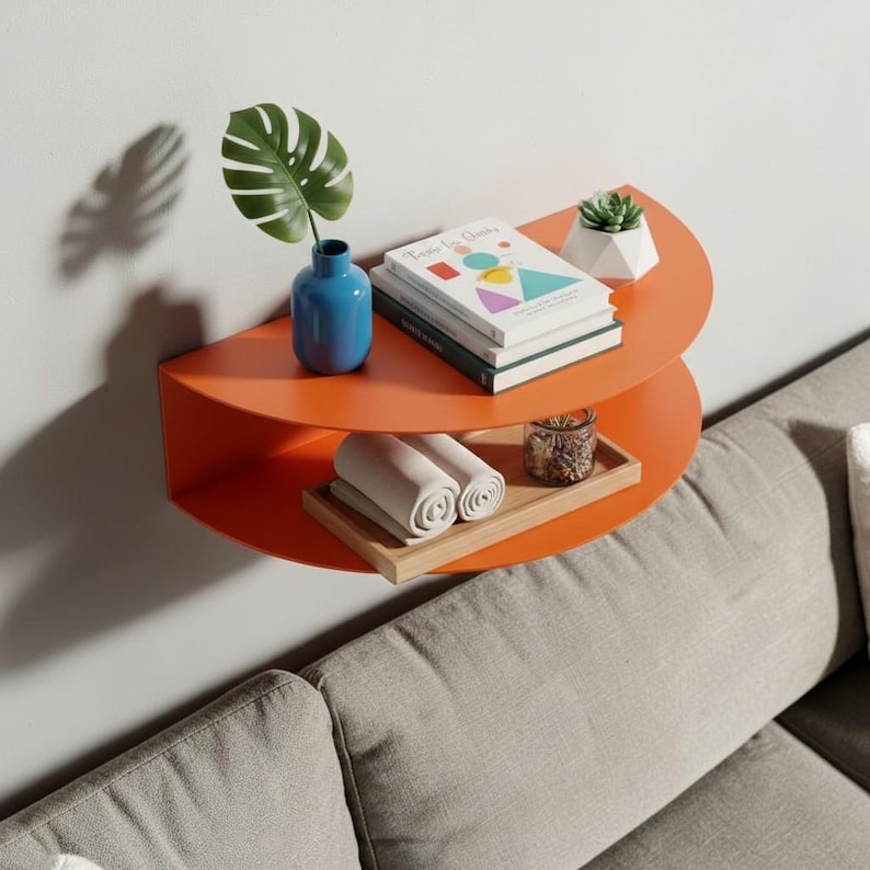 May include: An orange, semi-circular wall shelf with two levels. The top shelf holds a blue vase with a green leaf, books, and a small succulent. The bottom shelf displays rolled towels and a wooden tray with a glass jar.