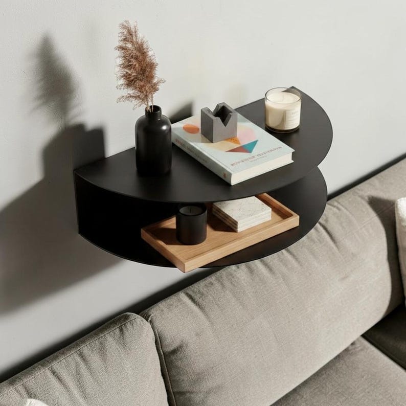 May include: A black, semi-circular wall shelf with two tiers. The top shelf holds a black vase with dried plants, a book, a gray geometric object, and a candle. The bottom shelf has a wooden tray and a black candle. The shelf is mounted on a white wall.