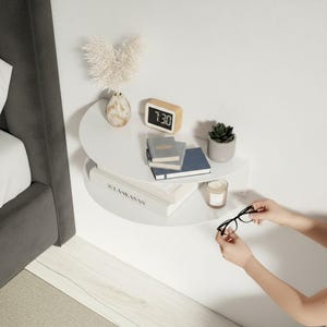 May include: A white, wall-mounted, semi-circular bedside table with a variety of items. A small wooden digital clock displays 7:30. There are books, a small potted plant, a candle, and a decorative vase. A person is holding a pair of black glasses.