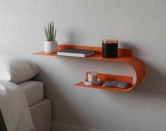 May include: A wall-mounted orange metal shelf with two tiers featuring a curved design. The top tier holds a white potted plant, a book, and a black canister. The bottom tier holds a small candle in a gray dish and a gray fabric eye mask.