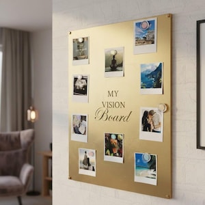 May include: A gold-coloured vision board with nine polaroid-style photos attached with clear magnets. The board has the words "MY VISION Board" in elegant script. The photos depict various scenes and images.