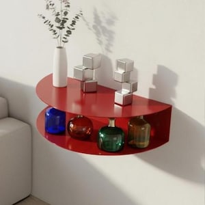 Peut inclure: Étagère murale rouge semi-circulaire à deux niveaux. L'étagère supérieure contient un vase blanc avec de la verdure et deux ensembles de sculptures de cubes argentés. L'étagère inférieure présente quatre bouteilles en verre colorées : bleu, rouge, vert et ambre.