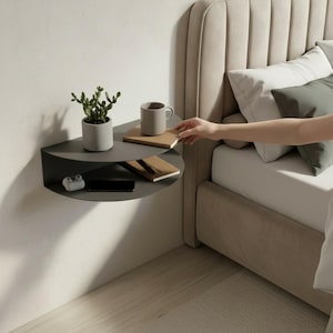 May include: A dark gray, semi-circular floating shelf mounted on a white wall. The shelf holds a small potted plant, a mug, a notepad, a smartphone, and a pair of wireless earbuds. A hand reaches towards the shelf from a bed.