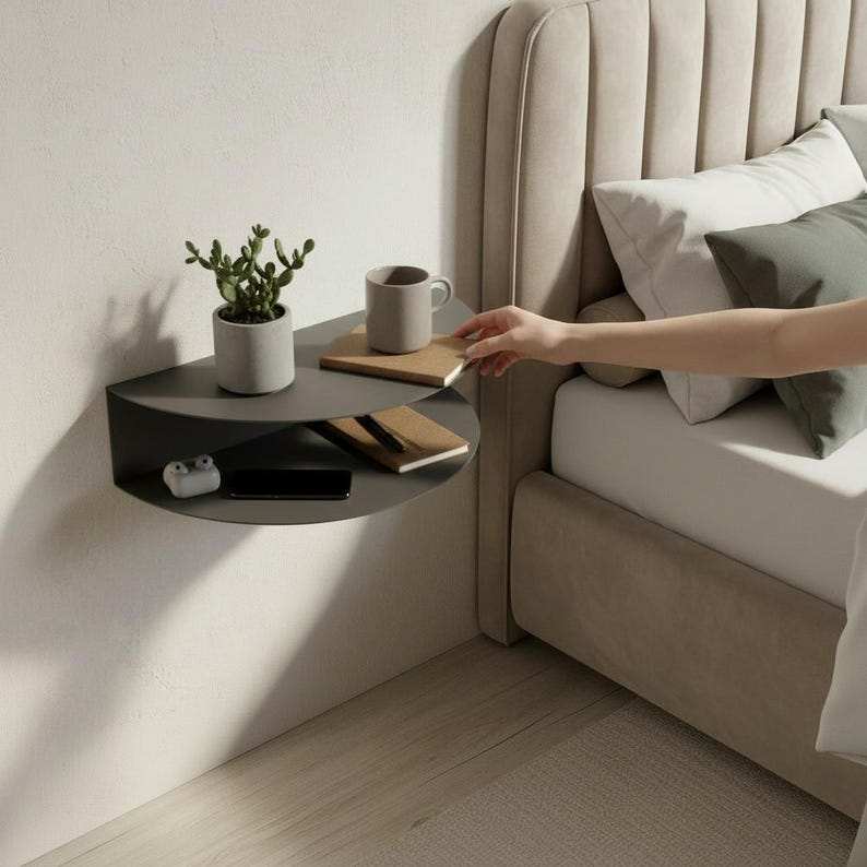 May include: A dark gray, semi-circular floating shelf mounted on a white wall. The shelf holds a small potted plant, a mug, a notepad, a smartphone, and a pair of wireless earbuds. A hand reaches towards the shelf from a bed.
