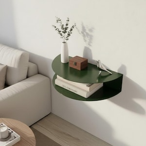 May include: A dark green, semi-circular wall shelf with two tiers. The top shelf holds a white vase with greenery, a small wooden box, and a decorative bird. The lower shelf displays two books. The shelf is mounted on a white wall.