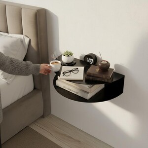 May include: A black, wall-mounted, semi-circular bedside shelf with books, a small succulent plant, a digital clock, and a cup of coffee. The shelf is mounted next to a bed with a beige headboard.