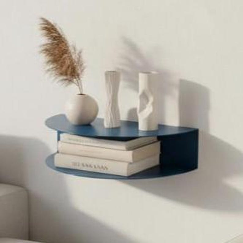 May include: A navy blue, semi-circular wall shelf with two levels. The top shelf holds a small white vase with dried pampas grass, and two white decorative vases. The bottom shelf displays a stack of books. The shelf is mounted on a white wall.