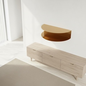 May include: A modern, gold-colored, semi-circular wall shelf is mounted on a white wall. Below, a light wood-toned cabinet with three drawers and tapered legs sits on a light-colored rug. The room has a minimalist design.