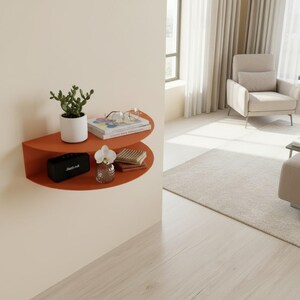 May include: A burnt orange, semi-circular wall shelf with various decorative items. A small potted plant, books, glasses, a black speaker, a small vase with a flower, and a stack of folded items are displayed on the shelf. The shelf is mounted on a cream-colored wall.