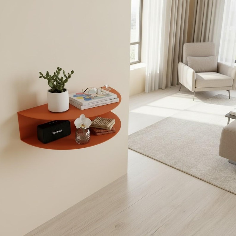 May include: A burnt orange, semi-circular wall shelf with various decorative items. A small potted plant, books, glasses, a black speaker, a small vase with a flower, and a stack of folded items are displayed on the shelf. The shelf is mounted on a cream-colored wall.