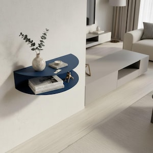 May include: A modern, navy blue wall shelf with a semi-circular design. The shelf holds a small vase with greenery, books, a candle, and a decorative brass frog. The shelf is mounted on a white wall in a contemporary living room setting.