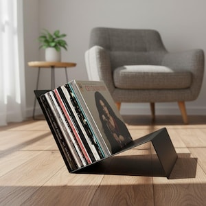 May include: A matte black metal vinyl record storage rack holding a collection of various music albums. The rack features an angled design that supports the records in an upright position, displaying the front cover of the first record.