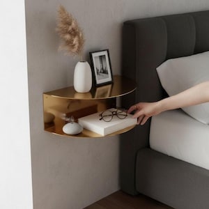 May include: A gold-coloured, semi-circular wall shelf with two tiers. The top shelf holds a white vase with dried flowers and a framed picture. The bottom shelf displays a book, eyeglasses, and a small decorative stone. The shelf is mounted on a grey wall.