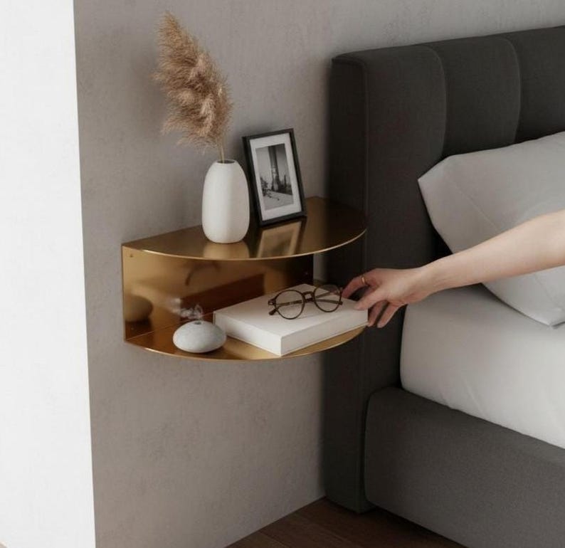 May include: A gold-colored, semi-circular wall shelf with two tiers. The top shelf holds a white vase with dried flowers and a framed picture. The bottom shelf displays a book, eyeglasses, and a small decorative stone. The shelf is mounted on a gray wall.