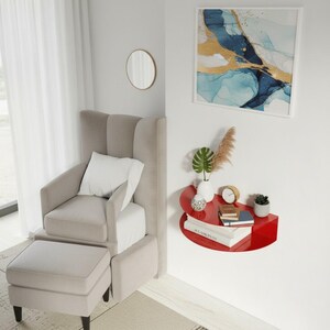 May include: A modern living room scene featuring a beige armchair with an ottoman, a red wall-mounted shelf, and an abstract painting with blue and gold accents. The shelf holds books, a clock, and decorative plants.