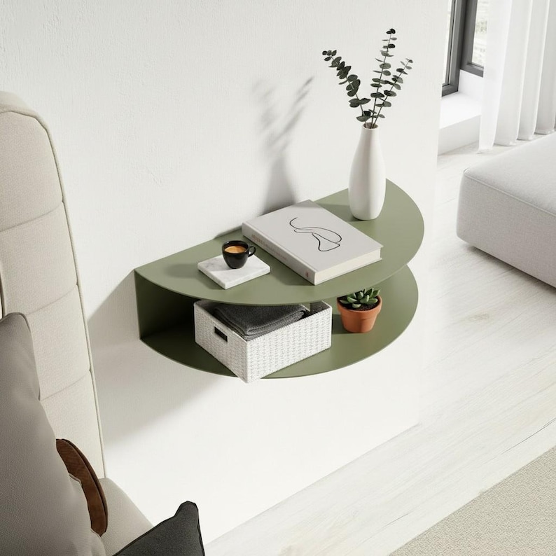 May include: A two-tiered, olive green, semi-circular wall shelf. The top shelf holds a book, a small cup of coffee on a white coaster, and a white vase with greenery. The bottom shelf has a small potted plant and a white woven basket.