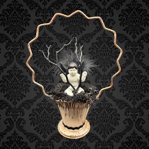 May include: A decorative arrangement featuring a small white cherub figurine with black wings and hair, nestled in a bed of dark moss inside a cream-colored, ornate vase. The arrangement is framed by a wavy, gold-toned metal border, set against a black damask wallpaper.